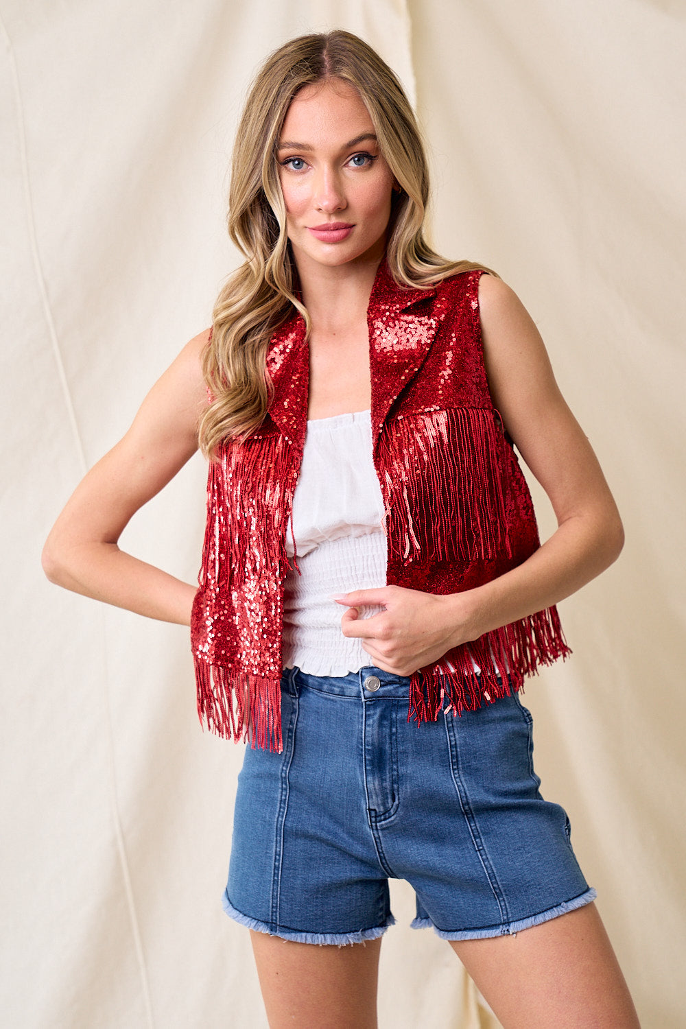 Woman wearing a red sequin vest over a white top and blue shorts on a beige background